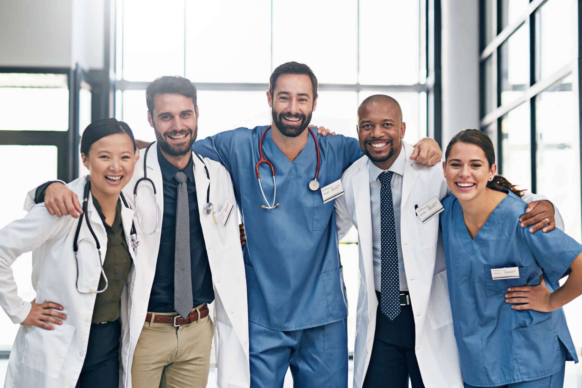 Team Medizin (1920 x 1280 px) Porträt einer Gruppe von 5 Ärzten, die im Krankenhaus stehen, sich umarmen und in die Kamera lächeln