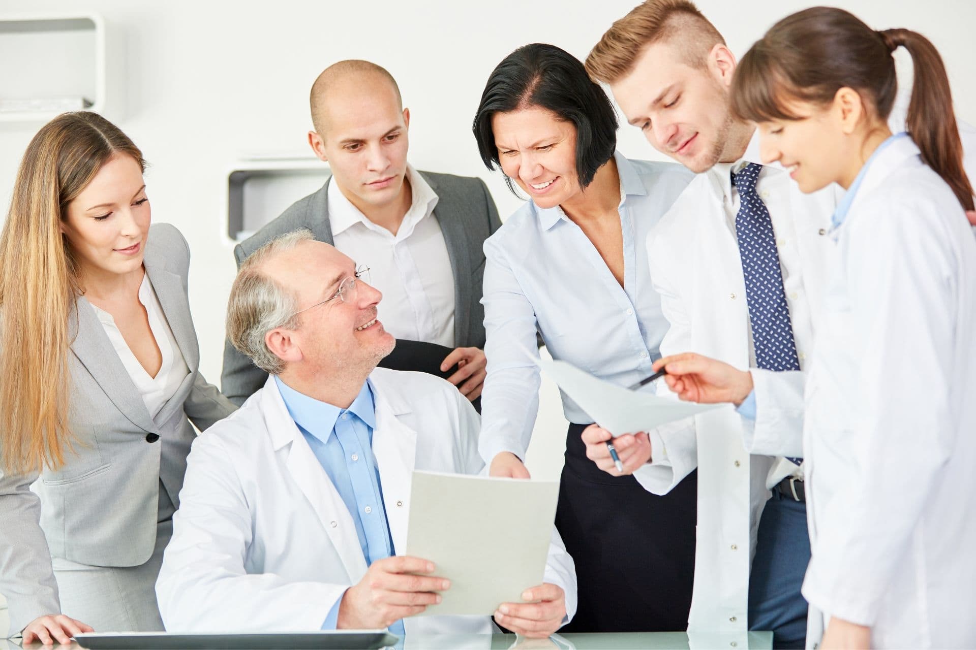 1 Arzt sitzend am Schreibtisch wird von 5 weiteren Mitarbeitern umringt, halten Papiere in der Hand, lächeln, strahlen Zuversicht aus